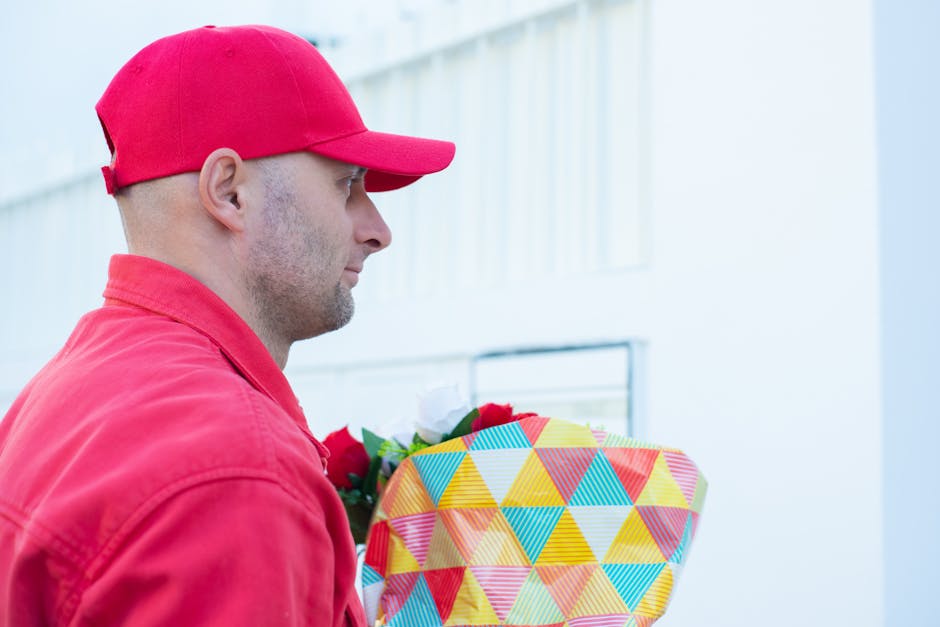 A delivery person wearing a red cap and matching red jacket is holding a vibrant floral gift wrapped in colorful geometric-patterned paper with shades of yellow, blue, green, and red. The person is in