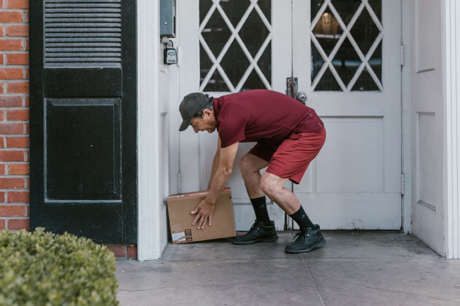 A delivery person wearing a burgundy t-shirt, matching shorts, black socks, and black work boots is bending down to pick up a small cardboard parcel from the doorstep of a house. The delivery is takin