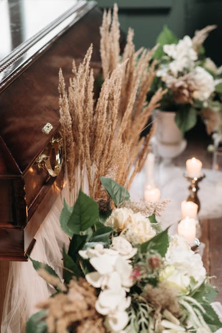 A floral arrangement placed on a polished wooden surface, featuring a mix of white and cream flowers with lush green leaves. Tall, beige pampas grass adds height and texture, creating an elegant, natu