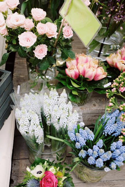 A display of various fresh floral arrangements including a bouquet of pale pink roses with dark green leaves, a cluster of pink tulips with green stems and leaves, and several vases of white and blue