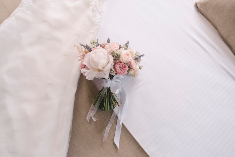 A delicate floral bouquet arranged with soft pink and white roses, peonies, and lavender sprigs, featuring lush green stems wrapped with a sheer white ribbon. The bouquet rests on a beige fabric surfa