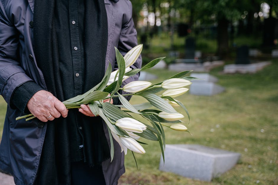 A person dressed in a dark jacket and shirt is holding a bouquet of white lilies with long green leaves, arranged with some flowers leaning slightly upward. The bouquet is wrapped loosely with no visi